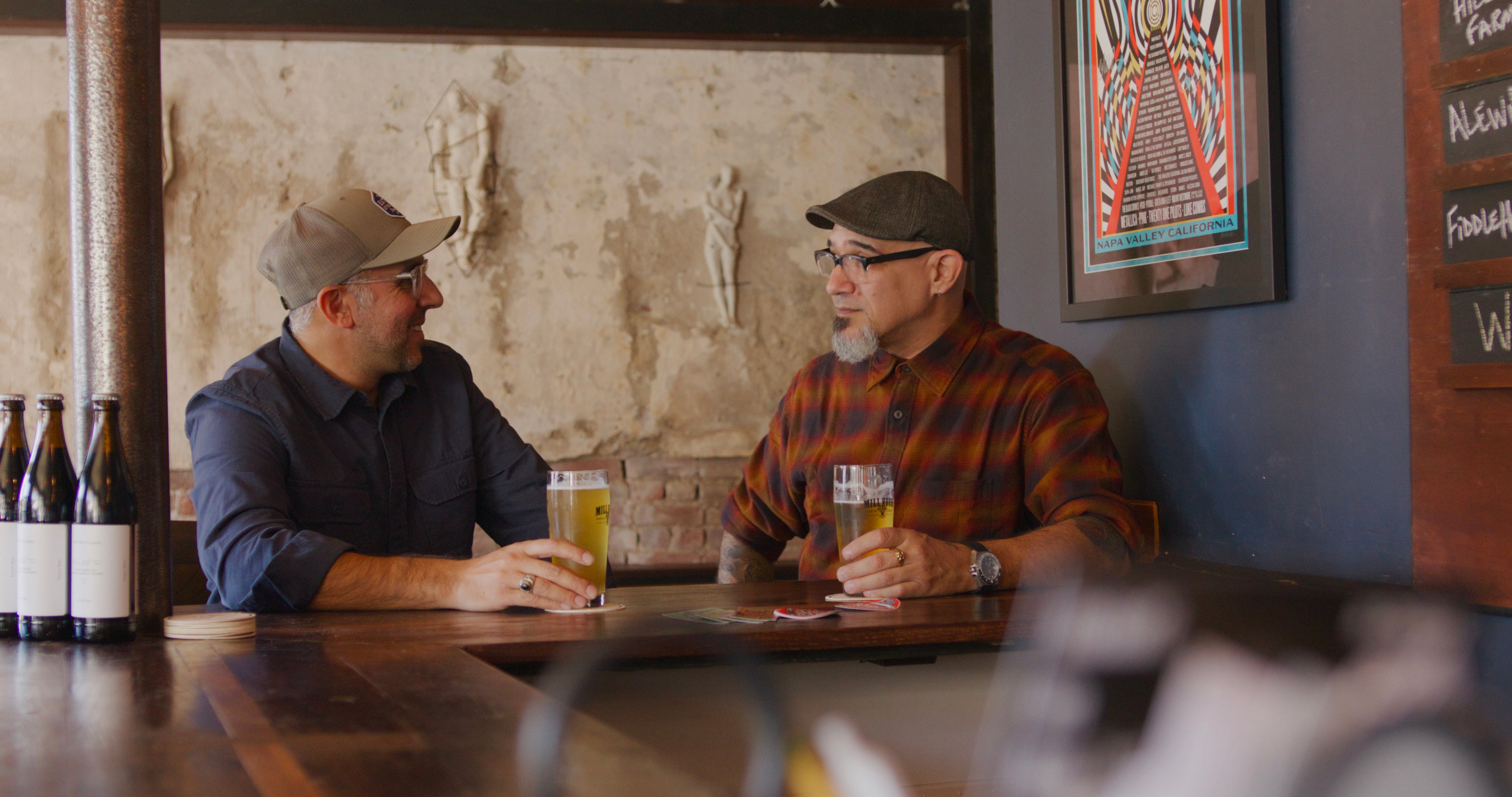 Two men sitting at a bar, each holding a beer, with bottles and a framed picture on the wall.